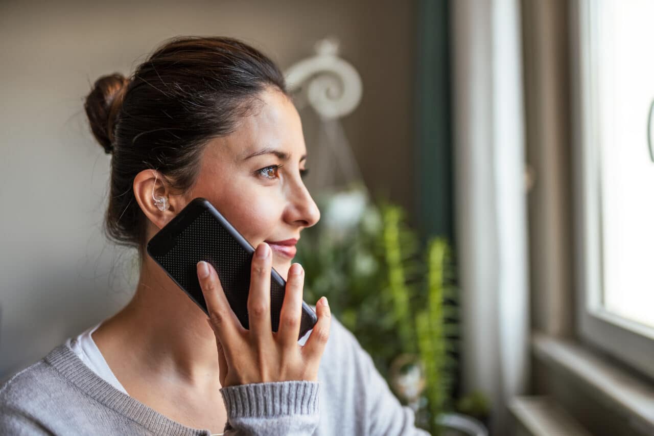 Young adult woman with a hearing aid speaks on her smartphone, looking out window.