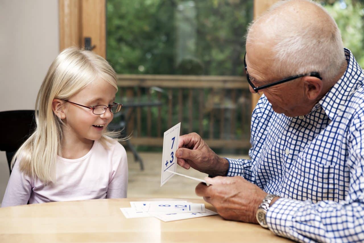A grandfather is holding an addition flashcard for his six year old granddaughter. Seated at a dining room table with patio door in the background, the girl is smiling and engaged in the activity.