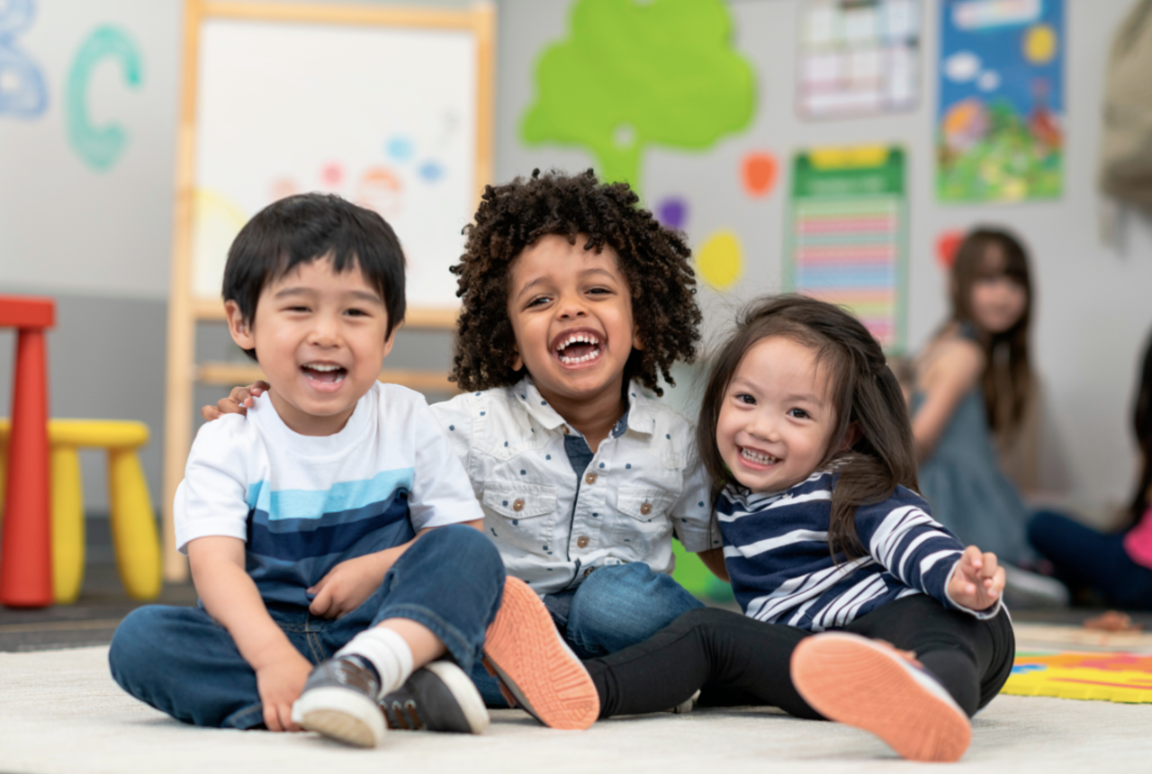 three toddlers are all smiles in their brightly colored preschool classroom